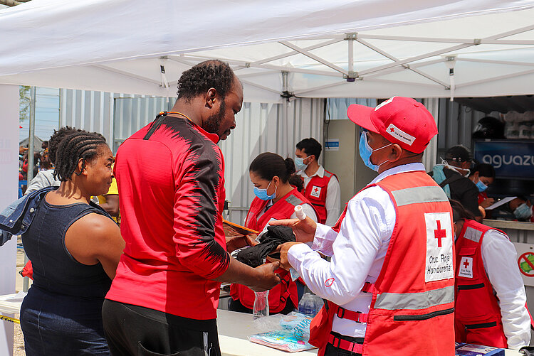 Personen erhalten Hilfe von Mitarbeitern des Honduranischen Roten Kreuzes unter einem weißen Zelt. Ein Mann in einem roten Oberteil nimmt Gegenstände aus den Händen eines Helfers im Rotkreuz-Outfit entgegen, während andere Personen warten und Gespräche führen. 
