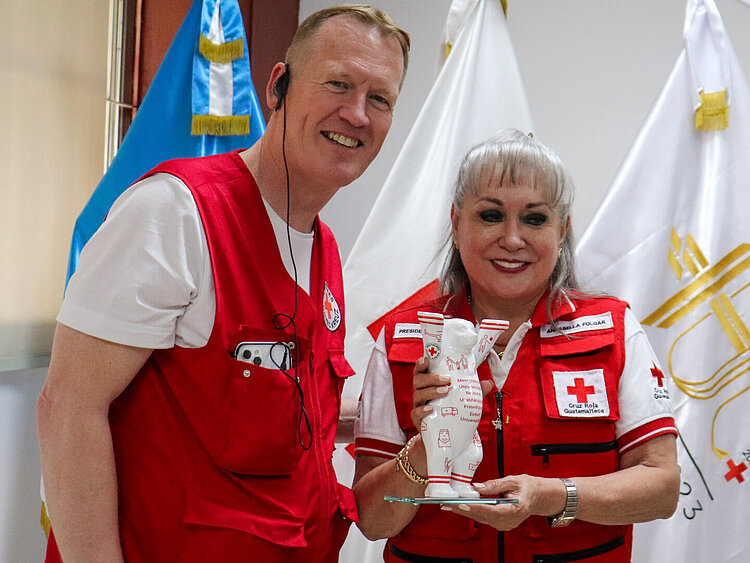 Christian Reuter und Annabella Folgar Bonilla, in Rote-Kreuz-Westen gekleidet, stehen lächelnd vor Flaggen. Die Frau hält einen Buddy Bear aus Porzellan mit Rote-Kreuz-Emblemen..
